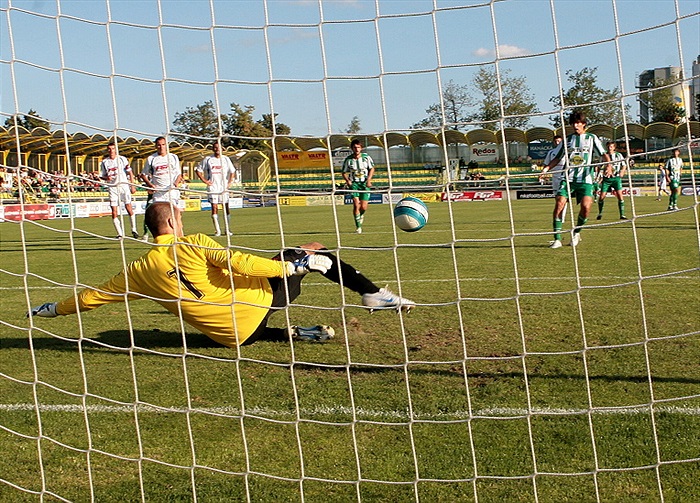 Brankář padá a míč nad jeho nohou míří do sítě.